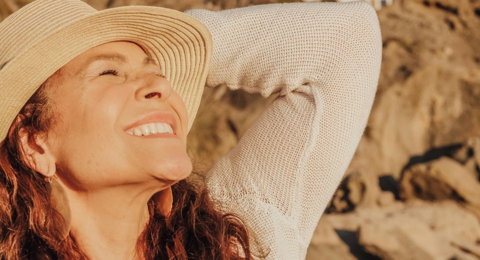 A joyful woman with a hat basks in the sunlight by the seaside, symbolizing relaxation and happiness.
