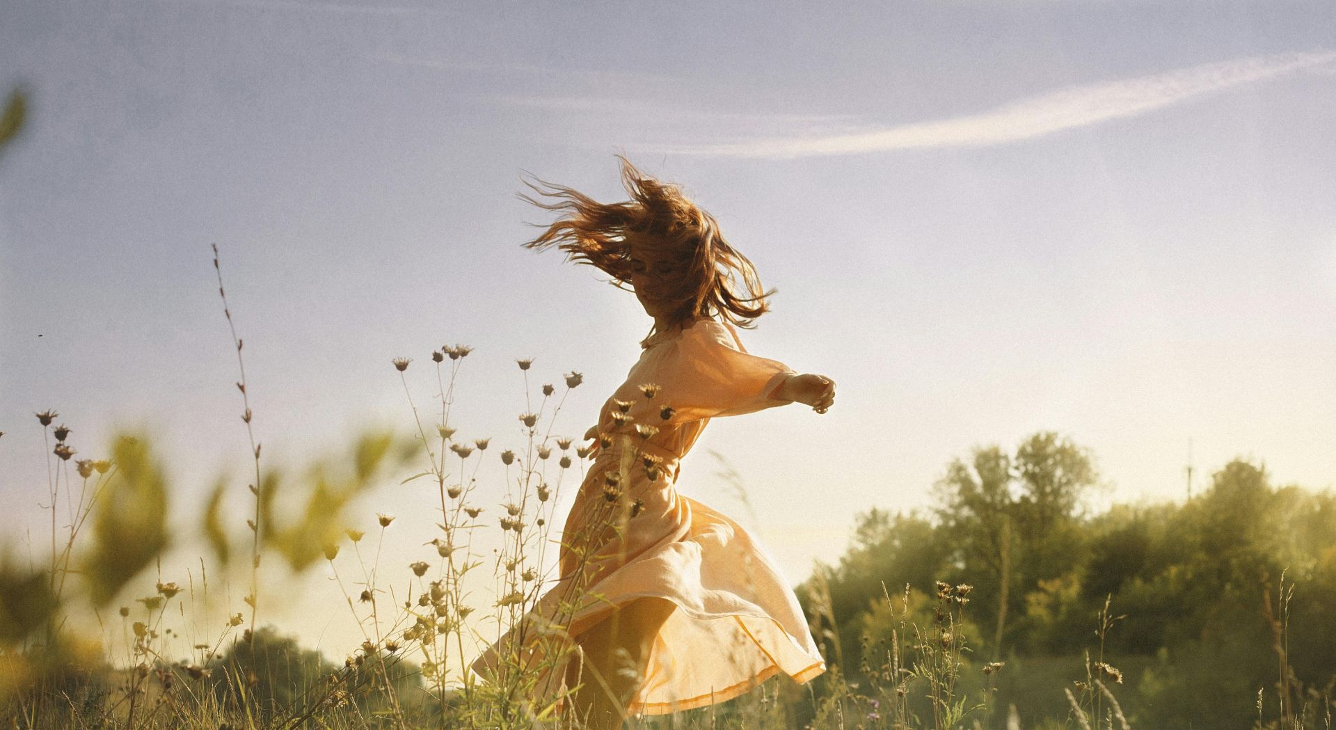 A carefree young woman in a dress dances joyfully in a sunlit meadow.